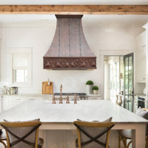 A handcrafted copper range hood above a kitchen stove, complementing the white cabinetry and tiled backsplash.